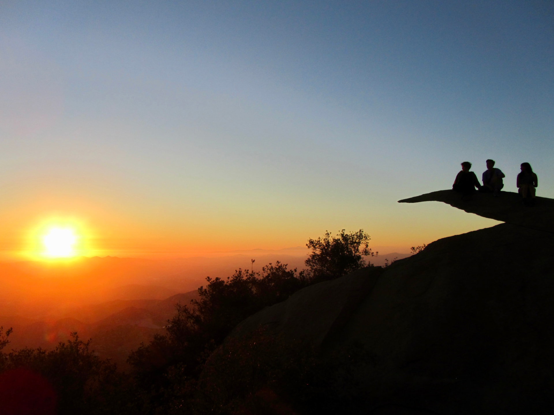 Three people sitting on a rock ledge watching the sunset over the hills